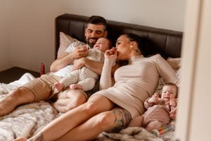 Parents relaxing on bed while feeding twin babies in casual home setting.