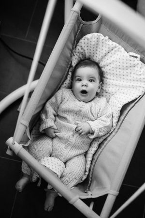Black and white photo of a baby in a knitted sweater lying in a baby bouncer with an excited expression.