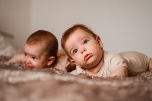 Two young infants lying on a soft blanket looking upward with curious expressions.
