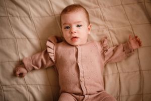 Baby wearing pink knitted outfit lying on beige bedding looking up at camera.