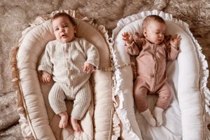 Two infants lying side by side in white baby loungers wearing soft knitted outfits.