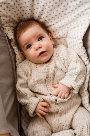 Baby wearing a cozy cream knit sweater lying on patterned fabric looking up with wonder.