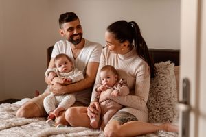Parents cuddling with twin babies on cozy bed in warm neutral tones.