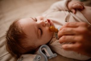 Baby with pacifier lying on bedding in warm lighting.