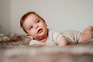 Baby in light colored onesie lying on plush carpet while gazing upward.
