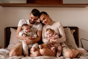 Family of four sitting together on bed with twin infants in matching outfits.