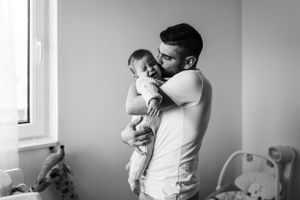 Black and white photo capturing tender moment between parent and infant by window.