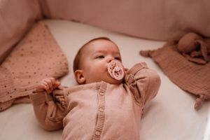 Baby wearing pink sweater lying on white bedding with pacifier.