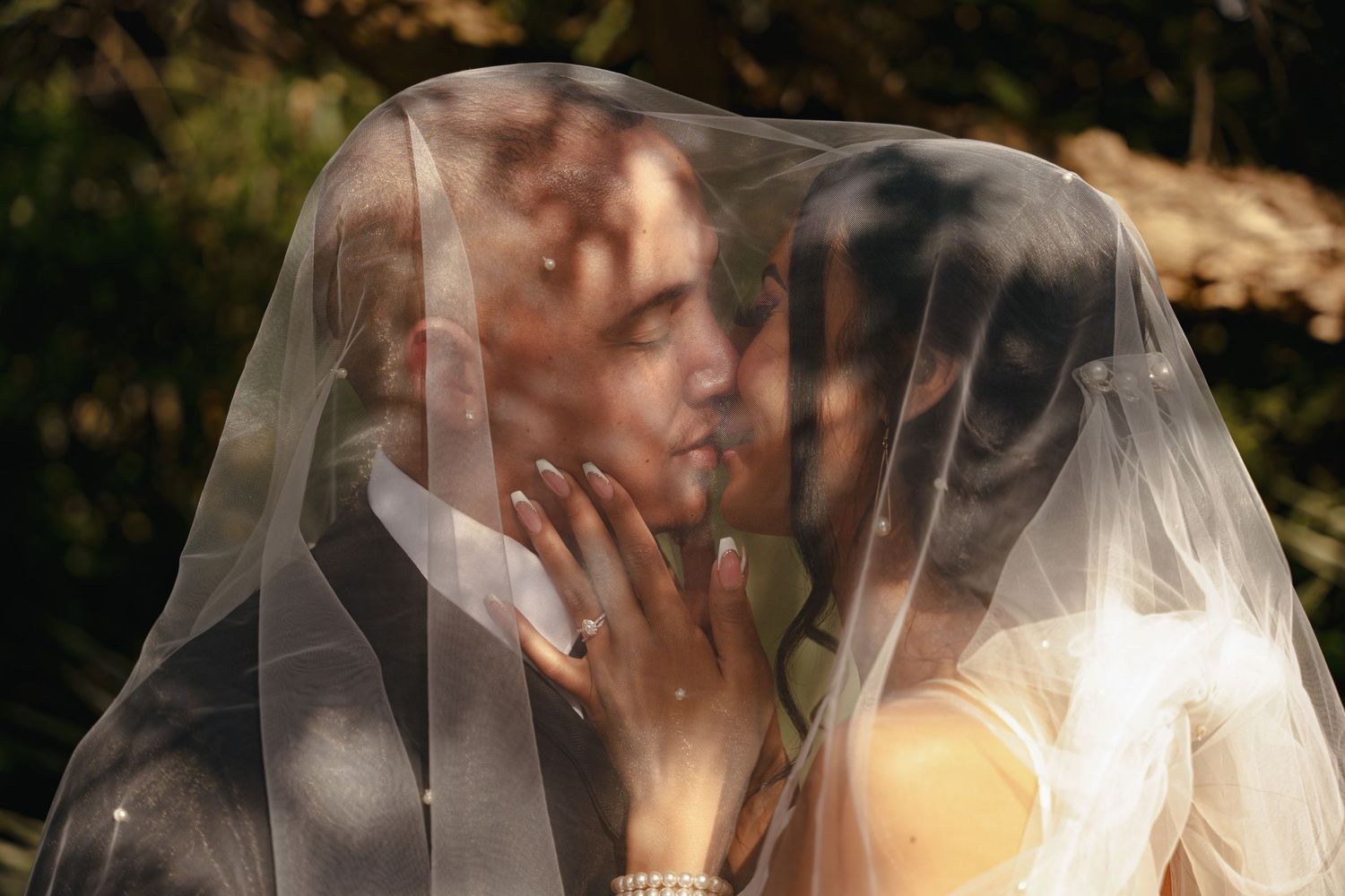 Romantic wedding photo of couple kissing under sheer veil with sunlight filtering through trees.