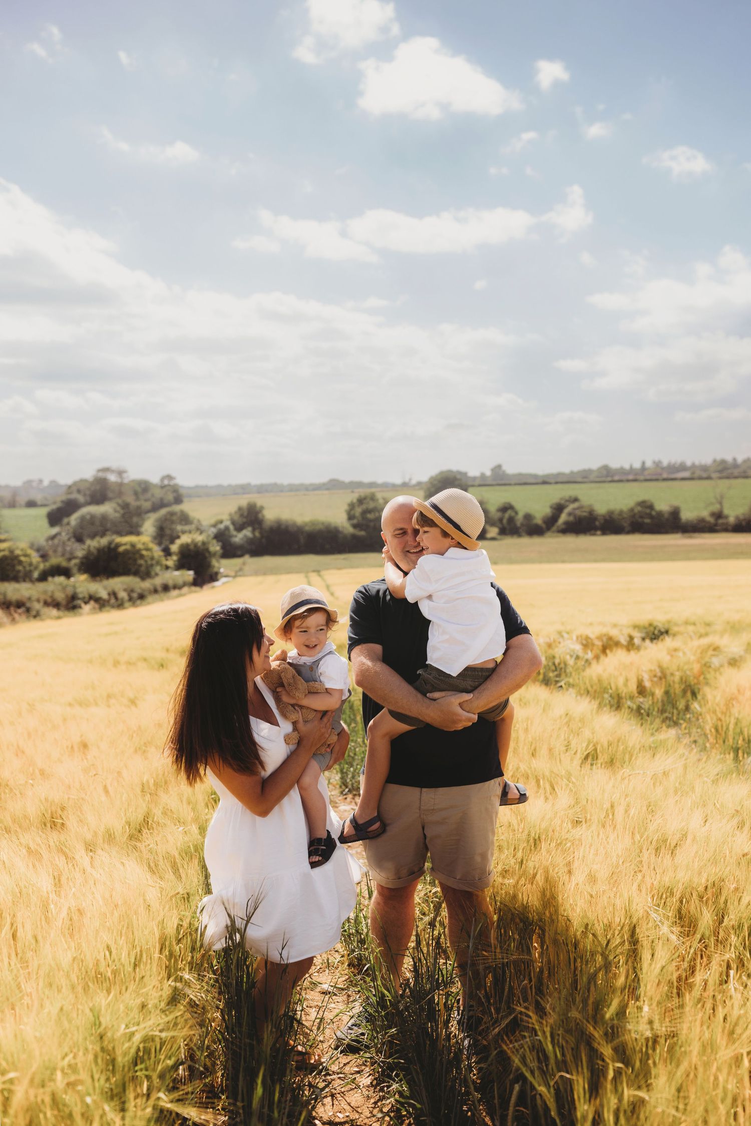Family standing together in golden wheat field under blue summer sky.