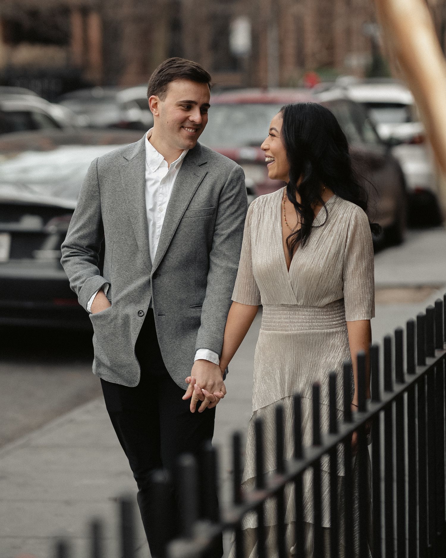 A stylish couple holding hands and laughing while walking along an iron fence in the city.