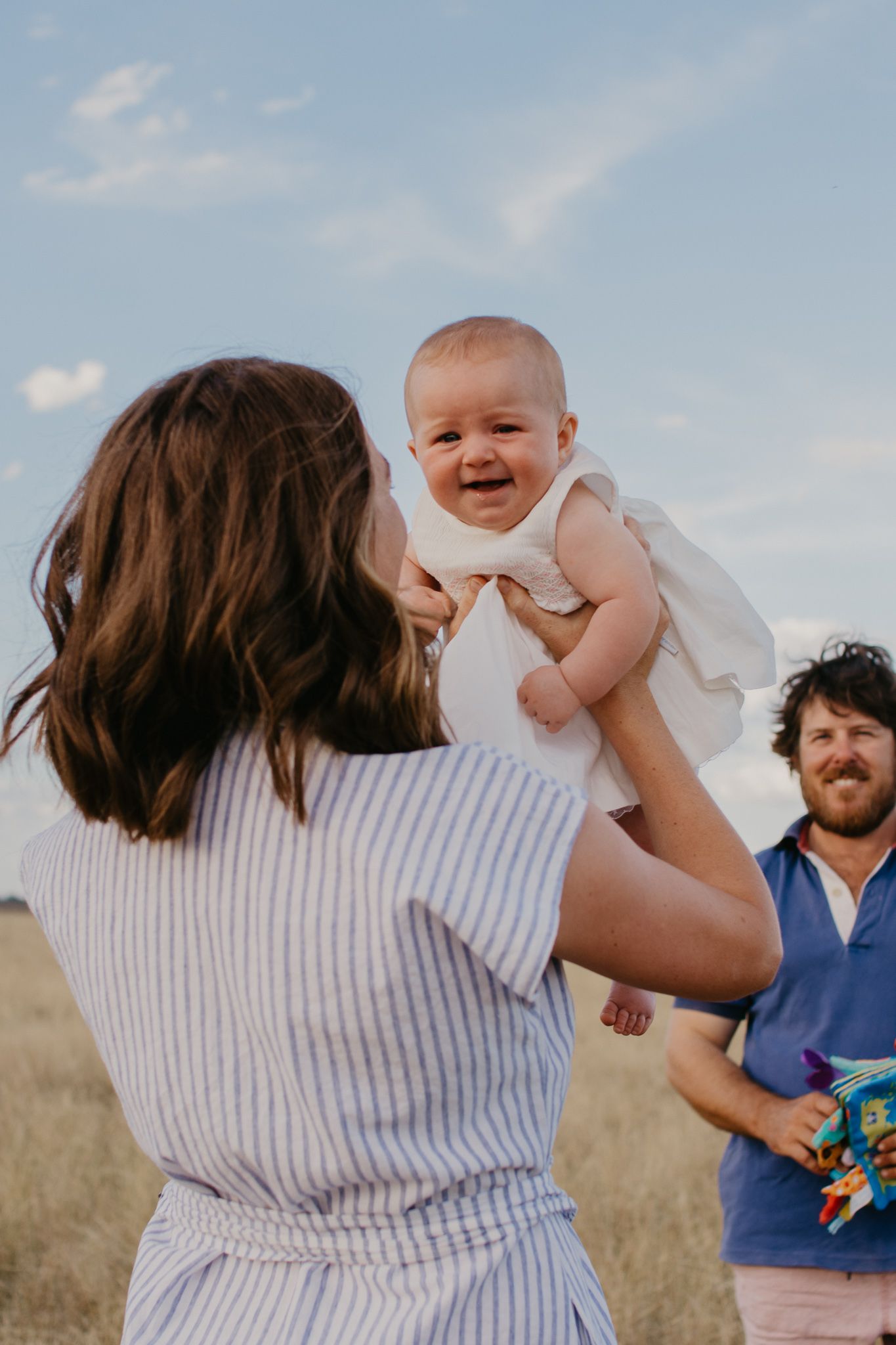 Mum holding baby high in the grass paddock and dad holding baby toys, looking at the both.