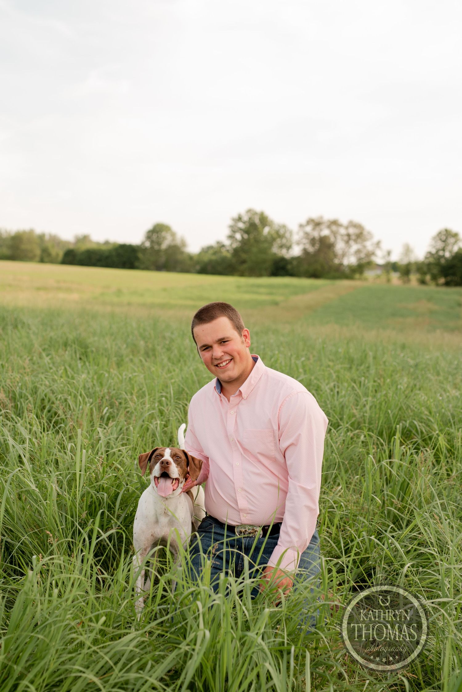 Person in pink shirt kneeling with brown and white dog in grassy field.