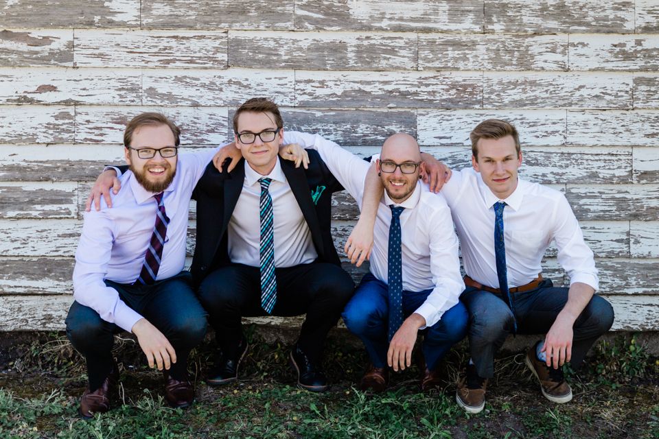 Four young men in dress shirts and ties pose together smiling against a rustic white wood wall.