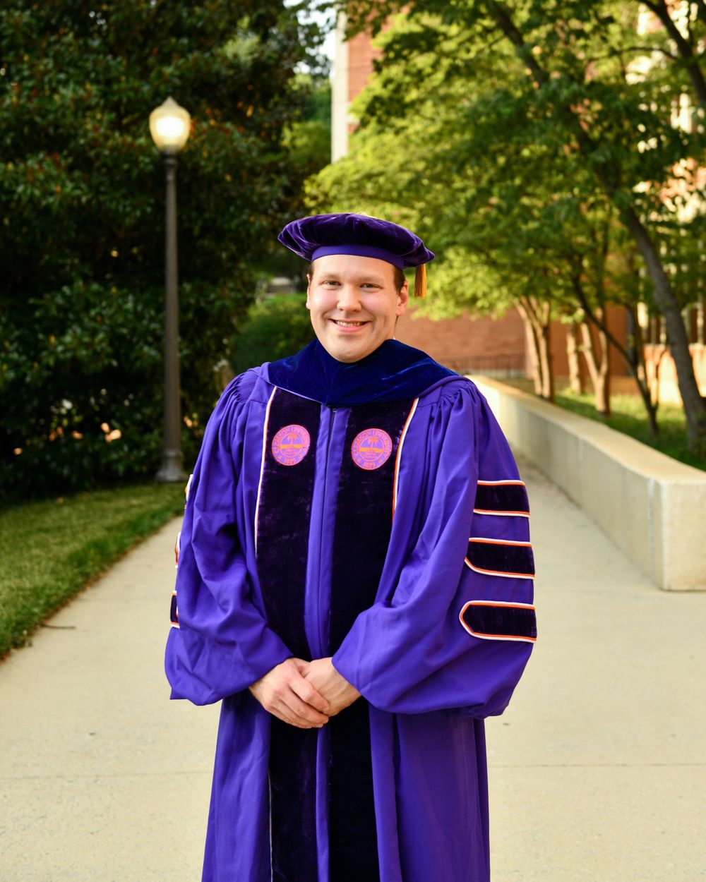 Graduate wearing purple doctoral regalia poses outdoors on campus walkway.