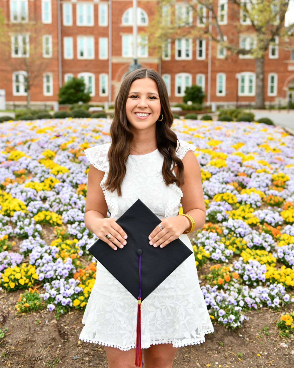 Graduate in white lace dress holding mortarboard in front of colorful flower garden.