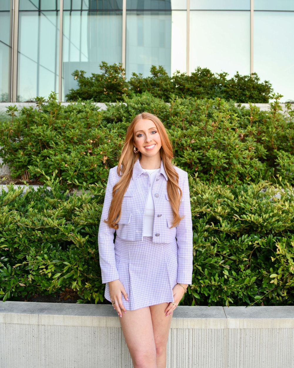 College senior poses in a purple outfit against a green hedge and glass building backdrop.