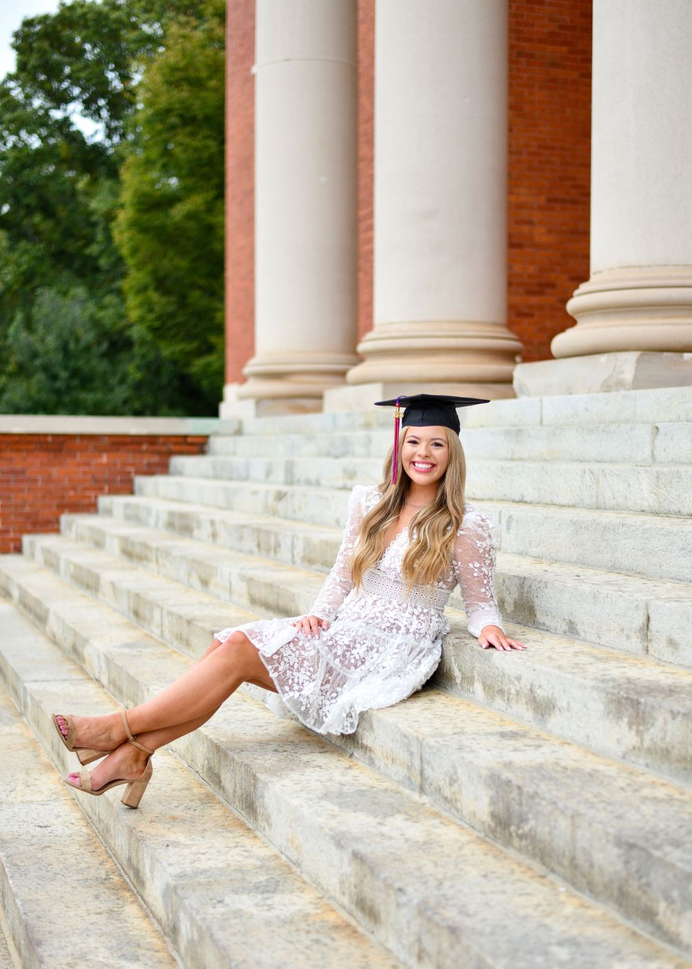 Graduate in a white lace dress sits on stone steps beside classical columns for a graduation photo.