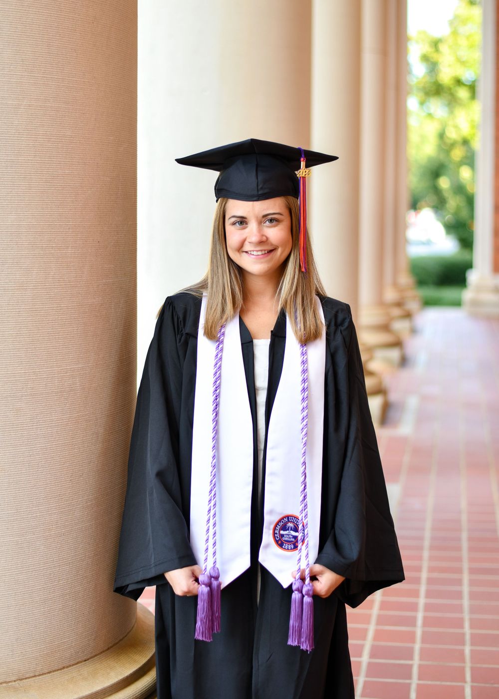 Graduate wearing cap and gown with purple tassels poses in front of brick columns.