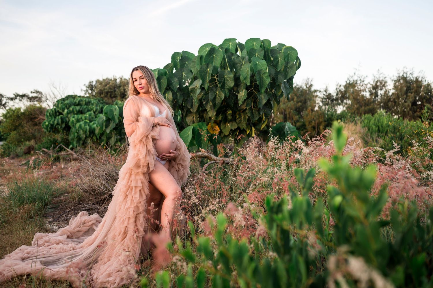 A maternity photoshoot in nature with a peach-colored flowing dress among green foliage.