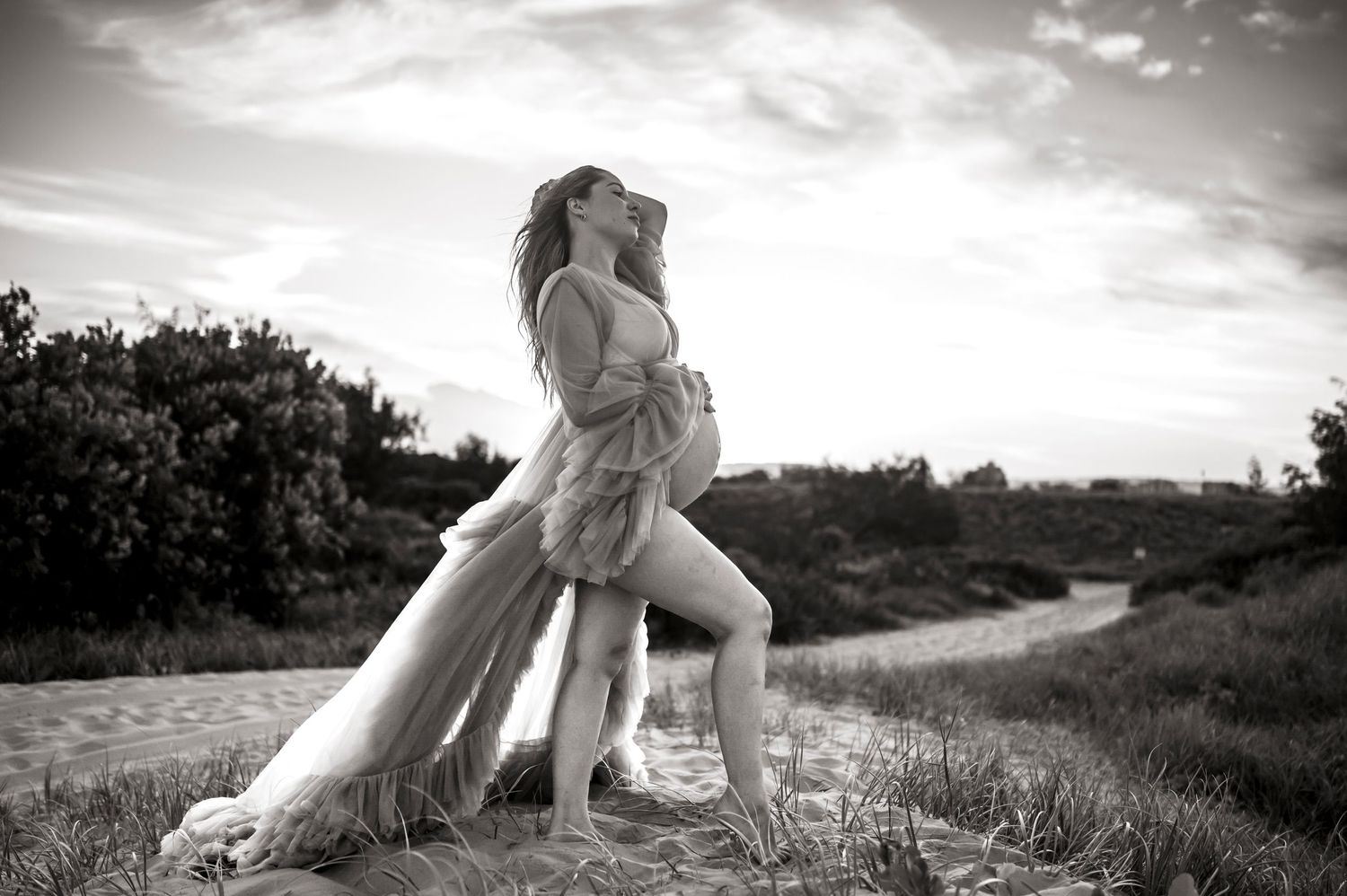 Black and white artistic maternity photograph on a hilltop path with flowing fabric.