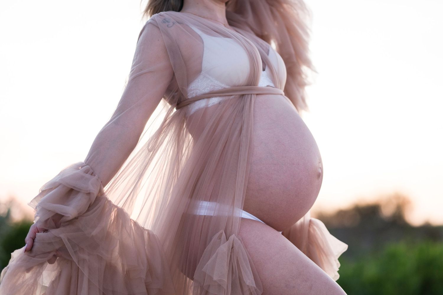 Close-up of a pregnant belly draped in delicate pink tulle fabric at sunset.