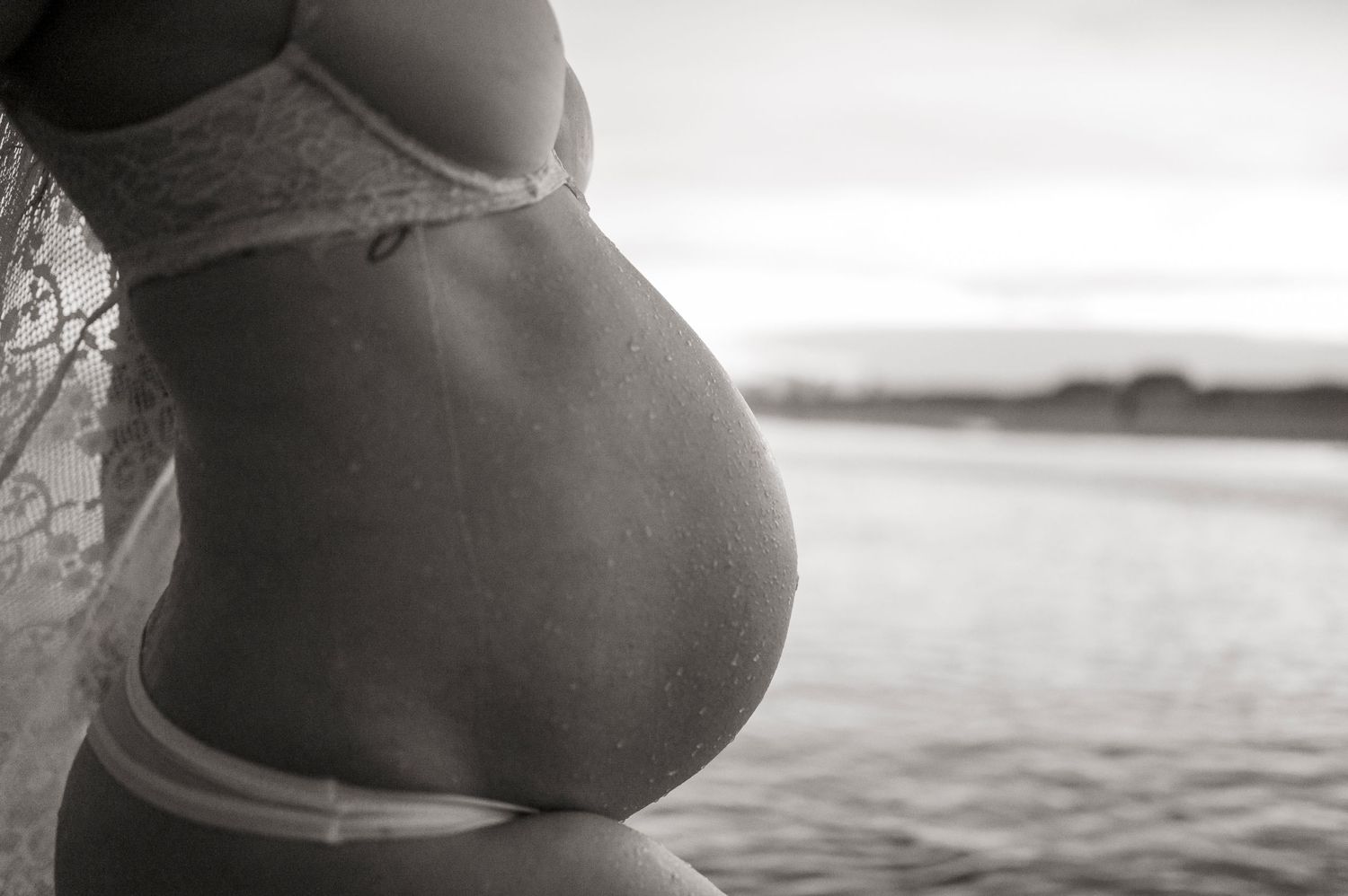 Side view of pregnant belly silhouetted against water in black and white.