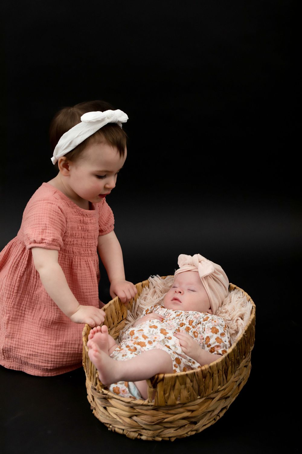 Two children with one standing beside woven basket containing sleeping baby.