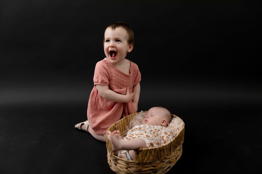 Child in coral dress laughing beside woven basket against dark background.