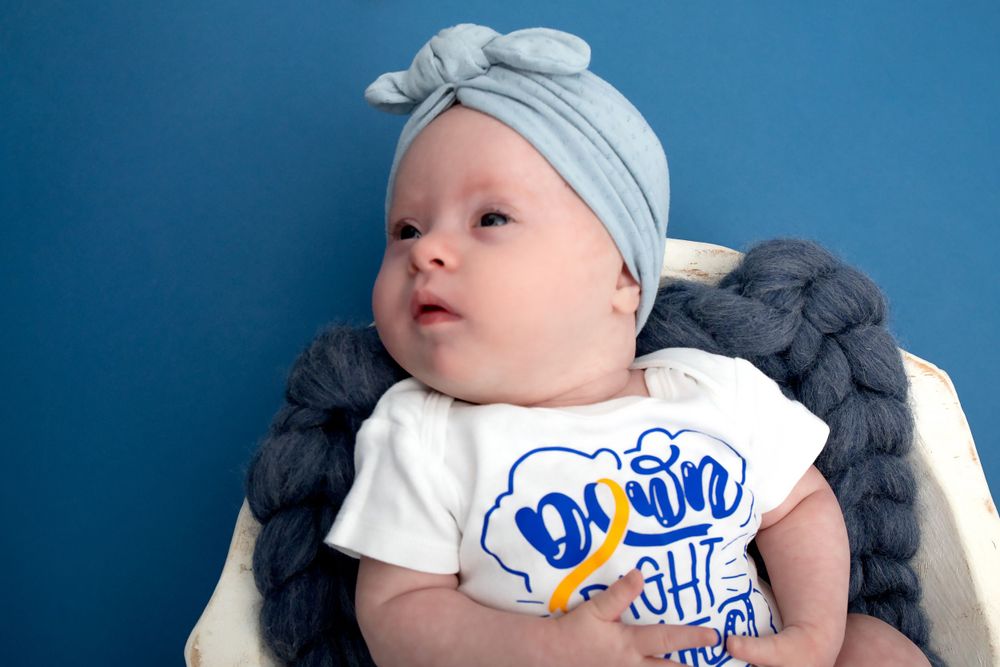 Baby in white graphic tee and blue bow headband against blue background.