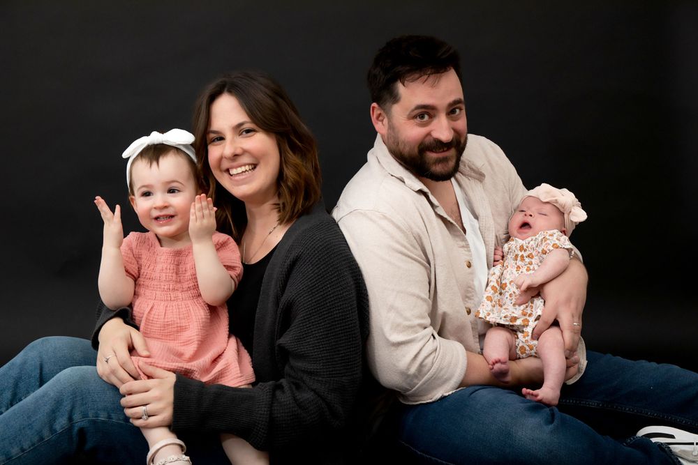 Studio family photo with parents and two children sitting together against black backdrop.