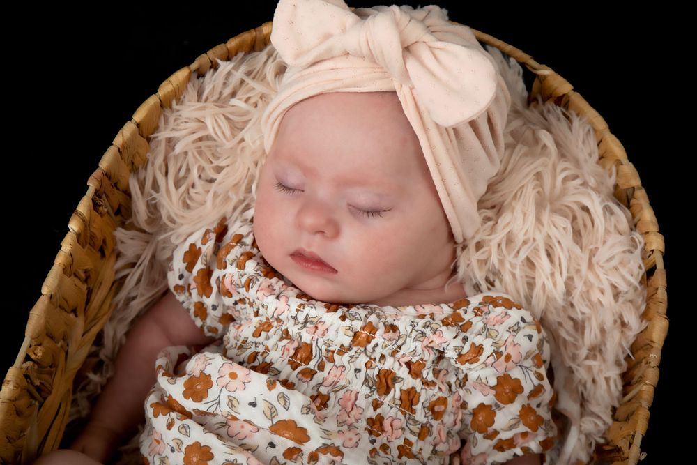 Sleeping baby wearing floral outfit with pink bow headband rests in basket with cream textured backdrop.