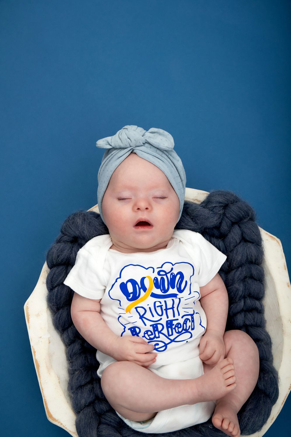 Baby wearing blue bow headband and graphic tee rests in basket with gray knit blanket.