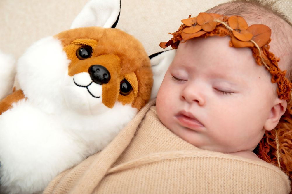 Sleeping baby with orange headband next to plush fox toy on cream background.