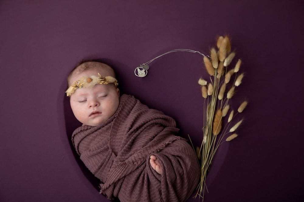 Swaddled sleeping baby with delicate headpiece on deep purple backdrop with wheat stalks.