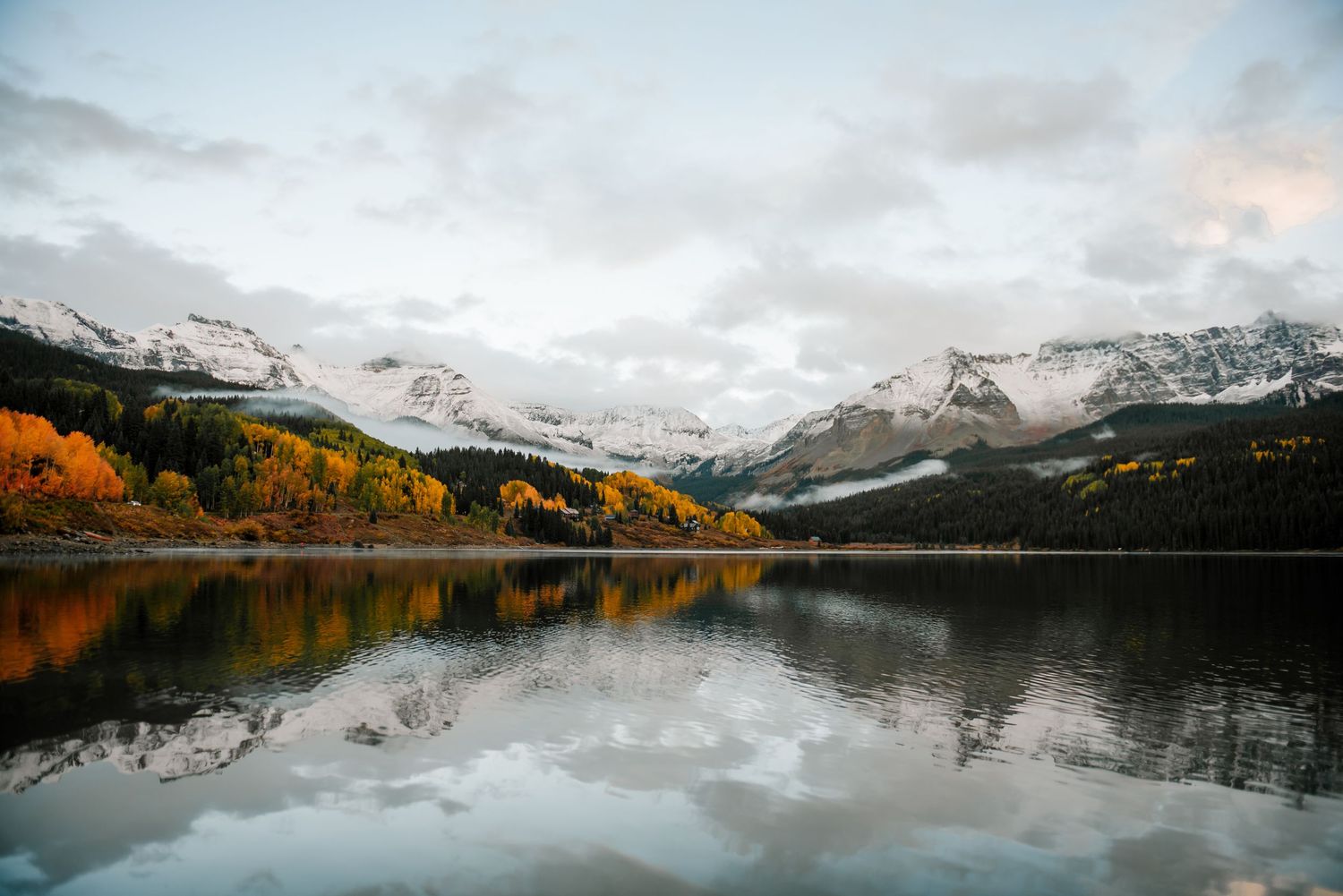Snow-capped mountains reflect in a serene lake surrounded by autumn foliage in a stunning mountain landscape.