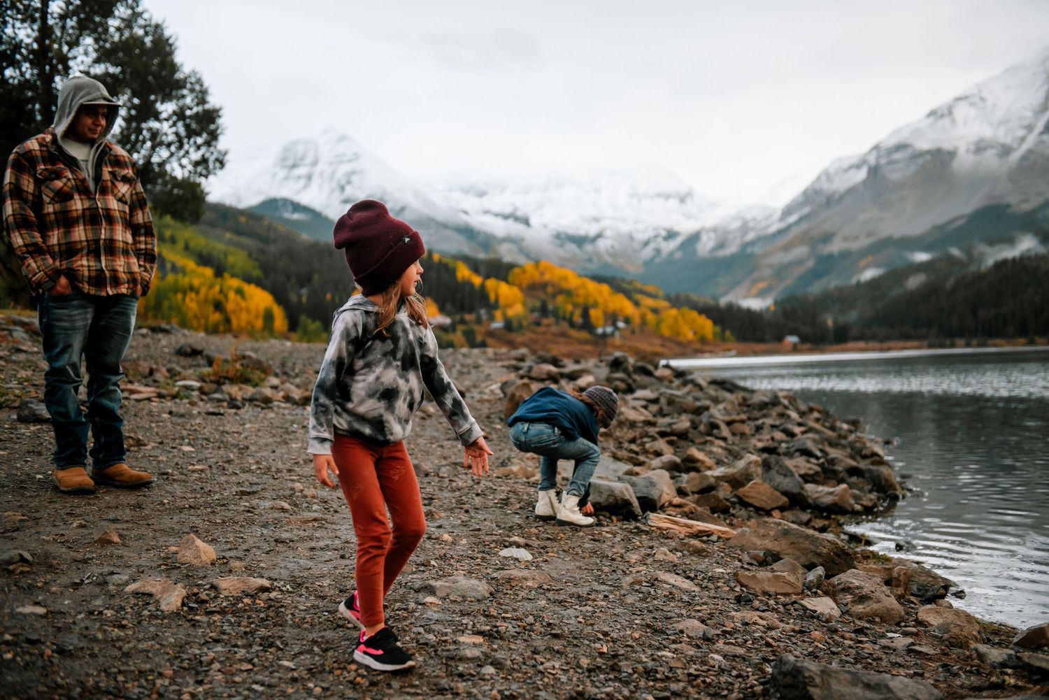 People relaxing on a rocky lakeshore with dramatic mountain scenery and autumn colors in the background.