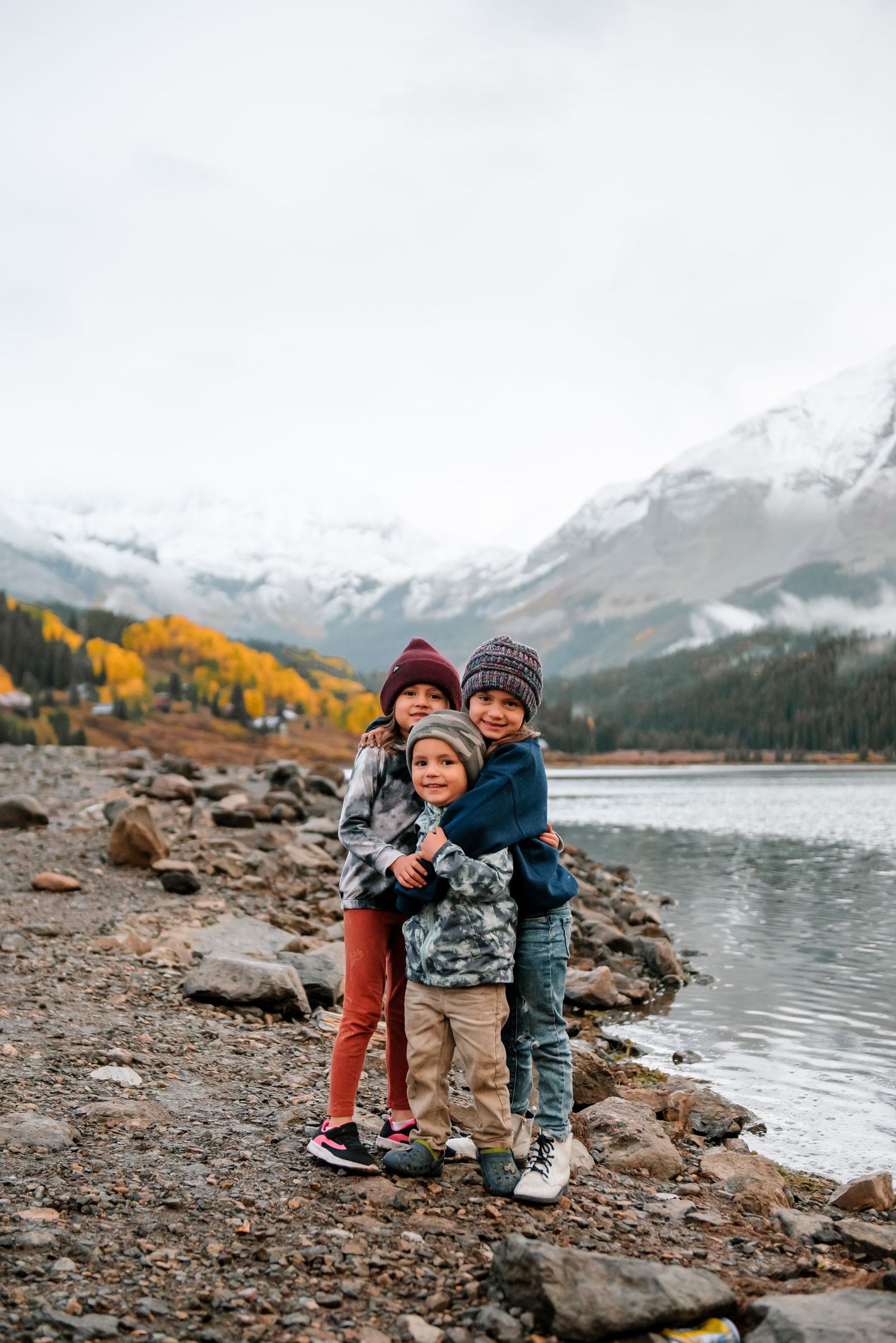 Children playing on rocky shoreline with mountain lake and snow-capped peaks in background.