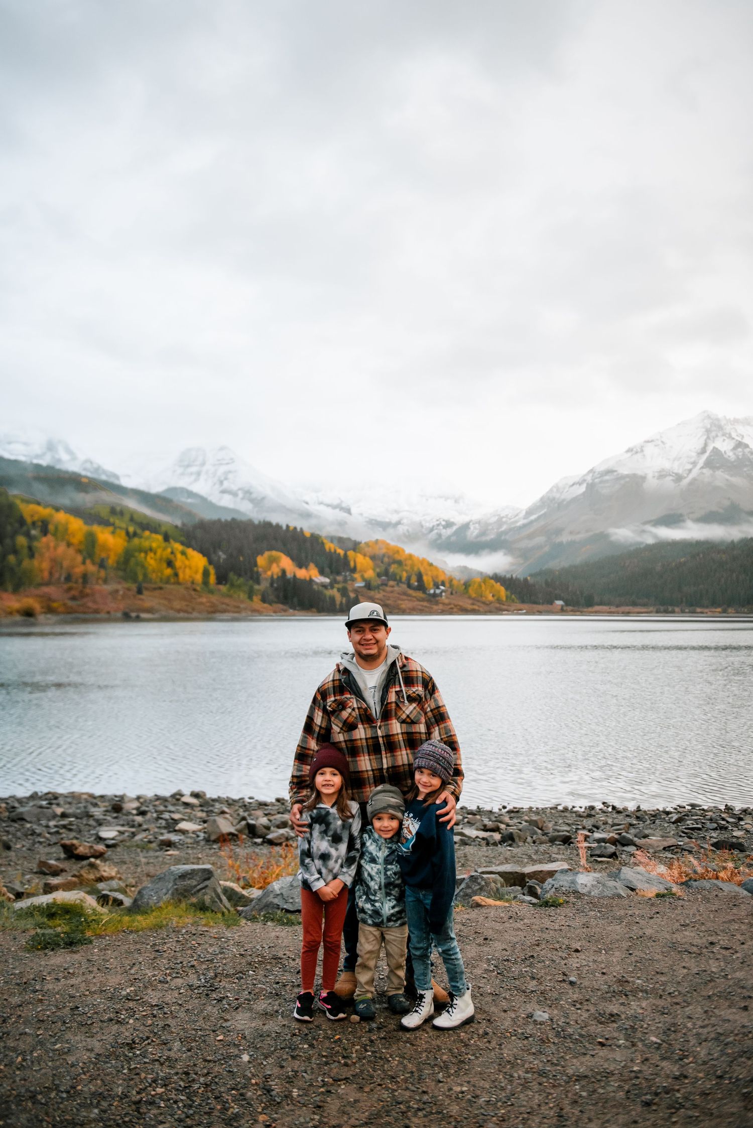 Family gathering on rocky shoreline with mountain lake and autumn foliage in background.