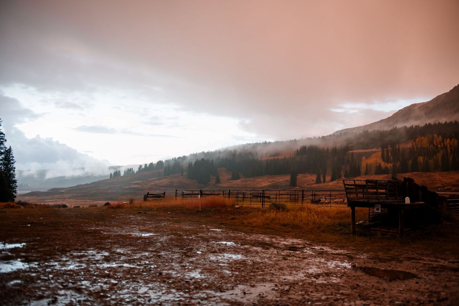 Moody mountain landscape with snow-dusted ground and rustic fencing under dramatic cloudy skies.