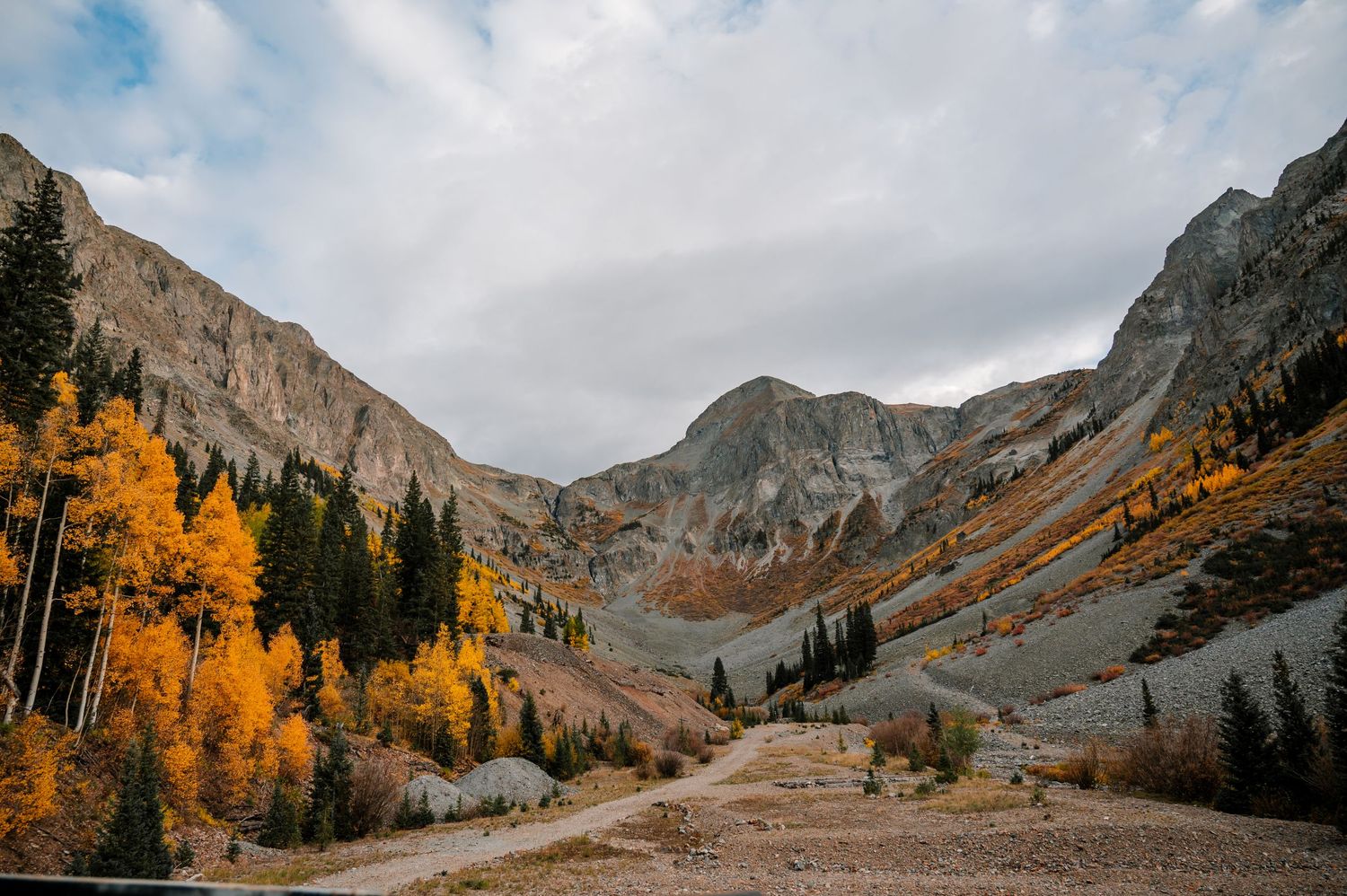 Dramatic mountain valley with golden aspens and evergreens under cloudy autumn skies.