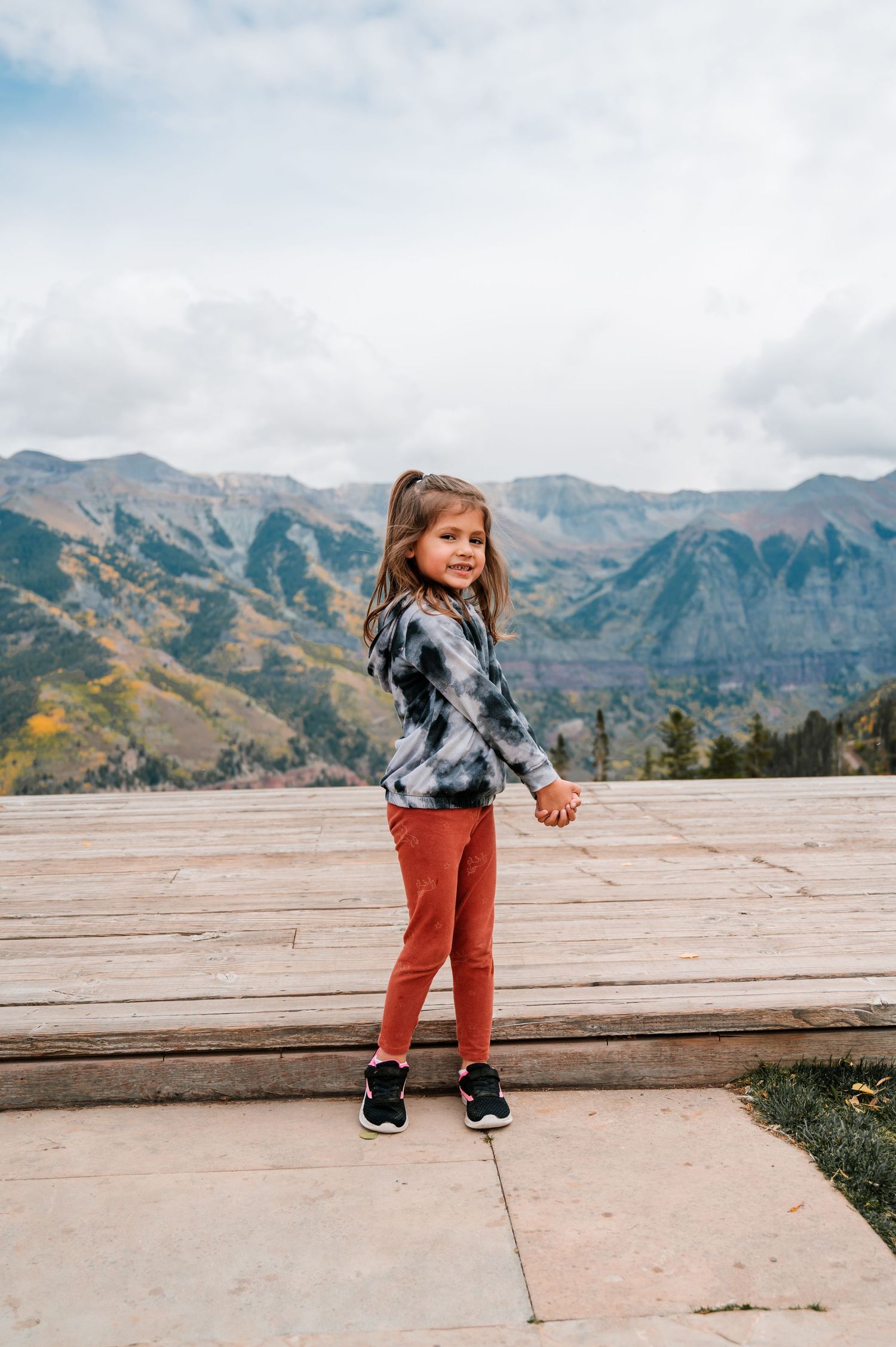 A toddler in red pants and plaid shirt dances on a wooden deck with mountains in the background.