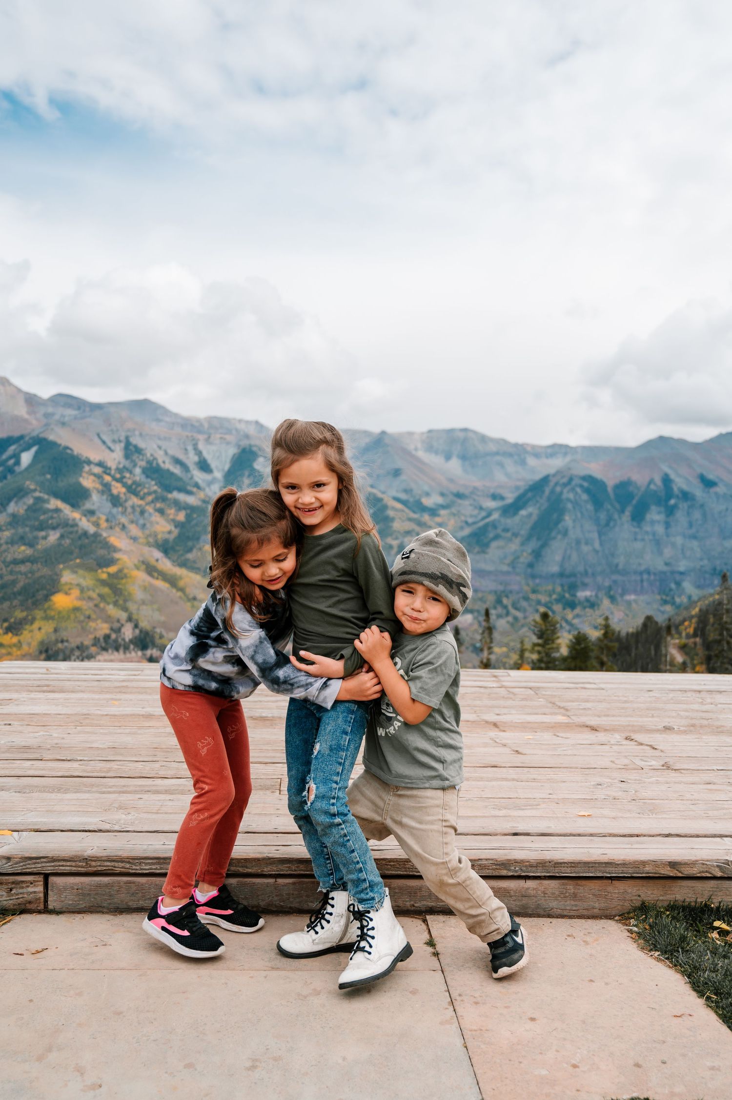 Three children play and laugh together on a wooden platform with scenic mountain views.