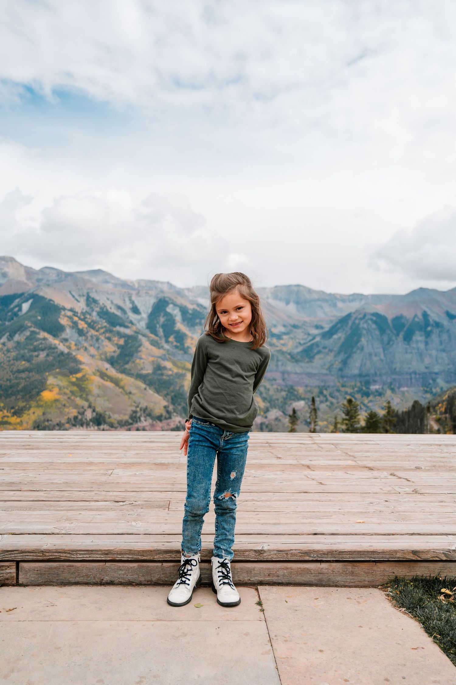 Child posing on outdoor wooden platform with scenic mountain views in background.