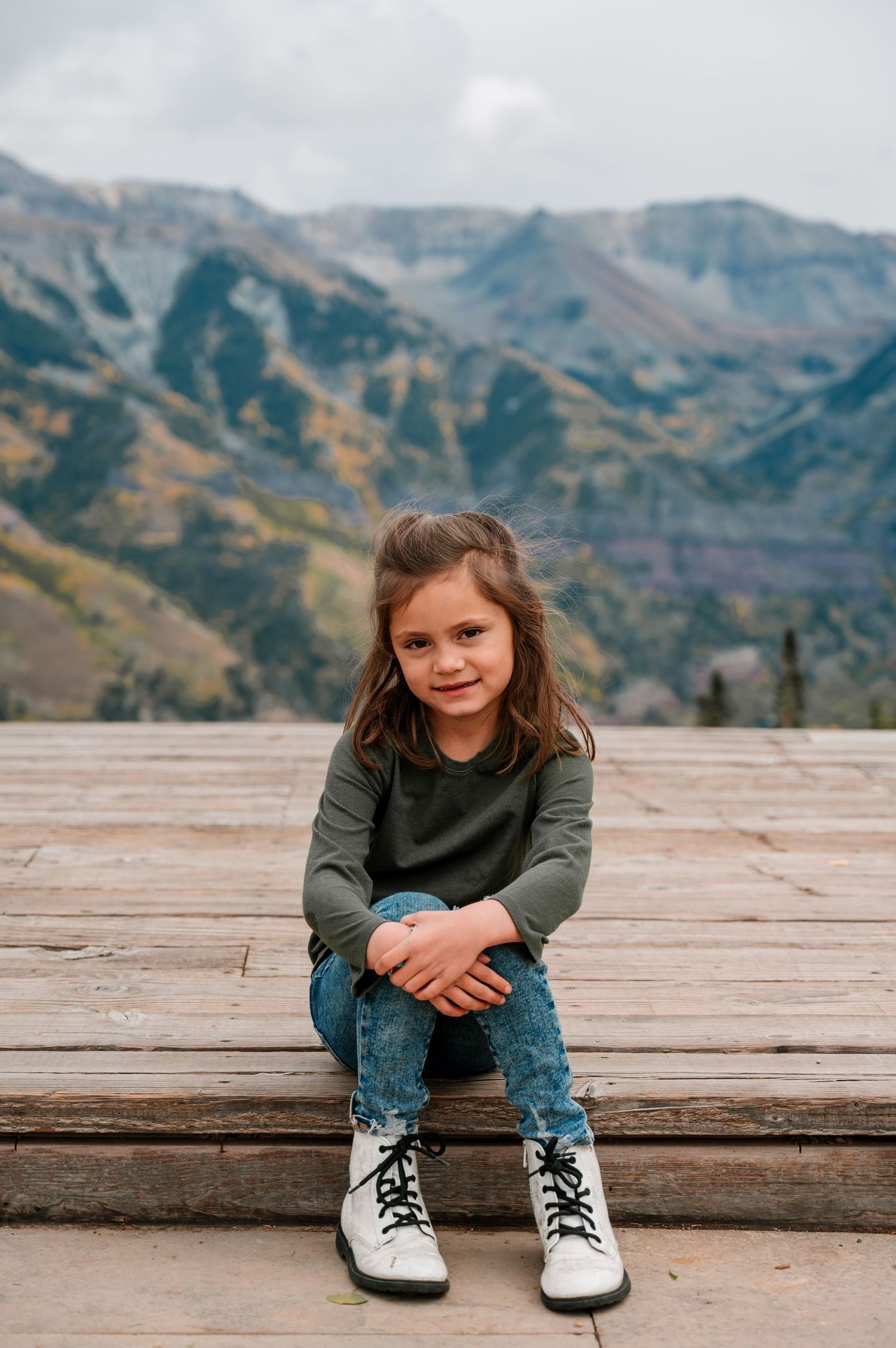 Child sitting on wooden deck with black and white sneakers against mountain backdrop in autumn.