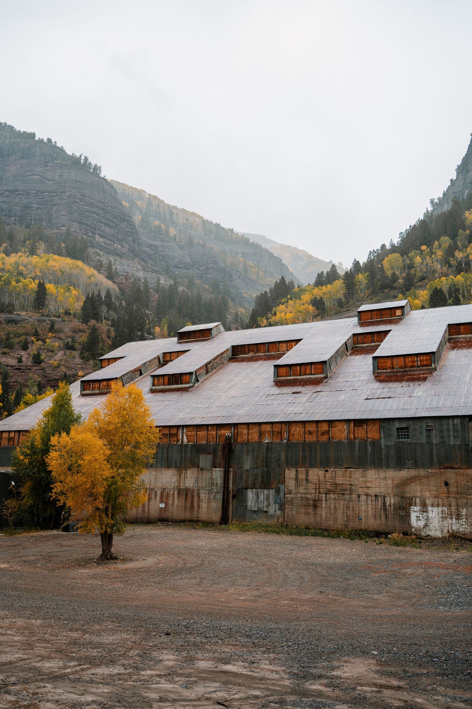 Row of industrial buildings with sloped roofs against mountain backdrop with fall foliage.