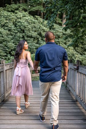 Couple walking hand in hand on wooden bridge surrounded by lush greenery.