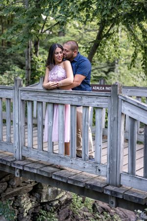 A romantic couple embraces on a wooden footbridge surrounded by lush greenery.