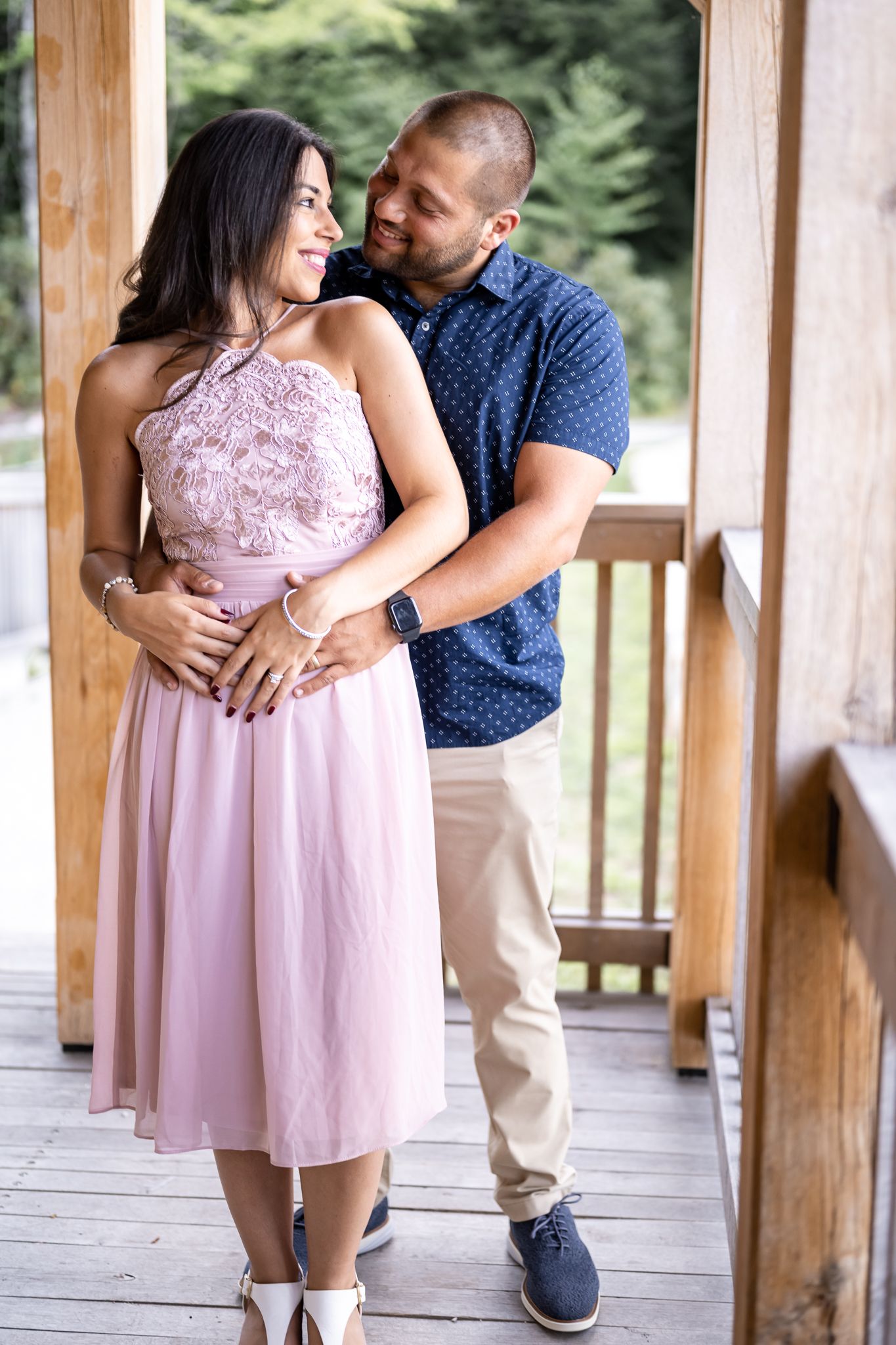 A couple shares an embrace on a wooden porch, with the person wearing a pink lace dress.