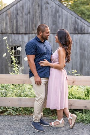 Engagement portrait in front of rustic barn with couple in coordinated casual outfits.
